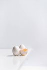 Alto ángulo de huevo entero y roto con yema en cáscara colocada en el borde de la mesa sobre fondo blanco en el estudio - foto de stock