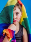 Menina bonito com lábios vermelhos e bandeira do arco-íris cobrindo metade da cabeça olhando para a câmera no fundo azul — Fotografia de Stock
