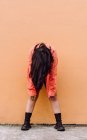Full body of young anonymous female covering face with long brown hair bending forward while standing against orange wall — Stock Photo