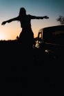 Back view of silhouette of unrecognizable woman with outstretched arms sitting on shoulders of man standing near car in nature on background of sundown sky — Stock Photo