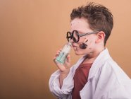 Химик в пластиковых очках с пятнами краски на лице, пахнущей жидкостью из бутылки на бежевом фоне — стоковое фото