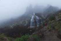 Turista ammirando cascate con corsi d'acqua veloci sul monte grezzo sotto il cielo nebbioso in autunno — Foto stock