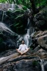 Friedlicher Mann sitzt in Padmasana mit Mudra-Händen und geschlossenen Augen, während er Yoga macht und auf nassen Felsen in der Nähe des Wasserfalls meditiert — Stockfoto