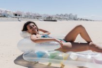 Vista laterale della donna felice in costume da bagno sdraiata sul materasso gonfiabile sulla spiaggia sabbiosa e prendere il sole nella giornata di sole durante le vacanze estive — Foto stock