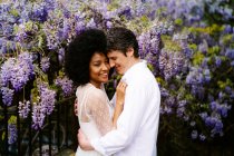 Вид збоку люблячої багаторасової пари, що обіймається в парку з квітучими фіолетовими квітами вістерії влітку — стокове фото