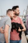 Vista lateral de alegres hombres homosexuales adultos con lindo perro mirando hacia otro lado en el puerto - foto de stock
