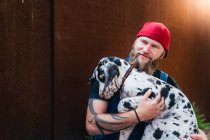 Happy bearded man with tattoos wearing denim overall and cap and sitting on cobblestone pavement hugging cute spotted Great Dane puppy — Stock Photo