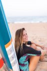 Enthousiaste surfeuse assise avec SUP board bleu sur le bord de mer sablonneux en été et regardant loin — Photo de stock