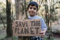 Criança étnica sorridente mostrando o título de Save The Planet na peça de papelão enquanto olha para a câmera na floresta — Fotografia de Stock