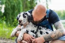 Pensive bald male with beard in casual outfit sitting with obedient spotted Great Dane puppy on lake with green trees on summer day — Stock Photo