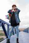 Maschio omosessuale elegante con bandiera LGBT colorata in piedi sul ponte e ascoltare musica in cuffia mentre guarda la fotocamera — Foto stock