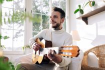 Contemplative male musician with tattoos playing classical guitar while sitting in armchair and looking away against window in house — Stock Photo