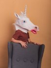 Anonymous kid in decorative unicorn mask with open mouth touching armchair on beige background — Stock Photo