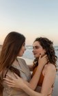 Молоді подружки обіймаються один з одним, стоячи на піщаному пляжі біля махаючого моря на світанку, дивлячись один на одного — стокове фото