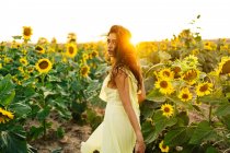 Gracieuse jeune femme hispanique en robe jaune élégante debout au milieu de tournesols en fleurs dans le champ de campagne dans la journée ensoleillée d'été en regardant la caméra — Photo de stock