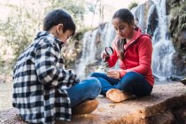 Ethnische Mädchen mit Bruder studiert Pflanzenblätter mit Lupe, während sie gegen Fluss und Kaskade sitzt — Stockfoto