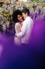 Vista laterale dell'amorevole coppia multirazziale che si abbraccia nel parco con fiori di glicine viola in fiore in estate — Foto stock