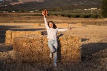 Zufriedene Frau mit Strohhut steht neben trockenem Heuhaufen auf Feld und blickt in Kamera — Stockfoto