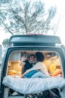 Jovem casal multirracial apaixonado tirando selfie no smartphone enquanto abraçando e beijando alegremente dentro van campista durante a viagem romântica juntos — Fotografia de Stock