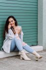 Charming female in stylish wear sitting near wall of building and leaning on hand while smiling and looking at camera — Stock Photo