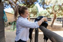 Side view of female jockey washing black saddle while preparing for horseback riding on farm in summer — Stock Photo