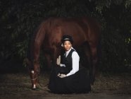 Señora adulta afroamericana confiada en ropa elegante y sombrero sentado con caballo marrón mientras mira lejos cerca de los árboles durante el día - foto de stock