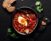 Top view of chakchouka with sunny side up egg in delicious tomato sauce with rye bread piece in bowl between fresh cilantro on dark background — Stock Photo