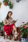 Zufriedene ethnische Frau in stylischem Outfit sitzt auf einem Hocker im Innenhof und liest Roman im Buch, während sie das Sommerwochenende genießt — Stockfoto