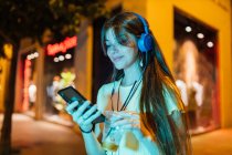 Заинтересованная женщина с напитками болтает по мобильному телефону, слушая музыку из гарнитуры в ночном городе — стоковое фото