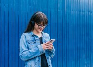 Mujer positiva en la navegación de auriculares en el teléfono inteligente mientras está de pie sobre fondo azul en la calle - foto de stock