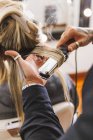 Anonymous male hairstylist using iron to curl blond locks of female customer during work in beauty salon — Stock Photo