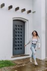 Mulher encantada em roupas da moda em pé perto da porta do edifício residencial e desfrutando de tempo chuvoso na cidade — Fotografia de Stock