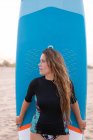 Surfista feminina feliz de pé com placa SUP azul na costa arenosa no verão e olhando para longe — Fotografia de Stock