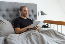 Pensive male sitting on soft bed in morning and reading interesting story in book after awakening — Stock Photo