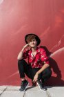 Trendy felice uomo omosessuale con unghie lunghe in camicia ornamentale guardando la fotocamera mentre accovacciato sul marciapiede contro parete — Foto stock