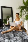 Mujer étnica contemplativa con netbook y vaso de agua descansando sobre una suave alfombra mientras mira hacia otro lado contra el espejo en casa - foto de stock
