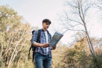 Low angle of male hiker with backpack navigating with paper map while standing in woods and looking away — Stock Photo
