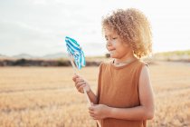 Вид сбоку на кудрявого волосатого ребенка, стоящего в сухой луже и играющего с игрушечной ветряной мельницей летом — стоковое фото