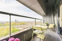 Стол со стульями расположен на просторном балконе современного жилого здания со стеклянным забором, смотрящим на городской пейзаж в летний день — стоковое фото