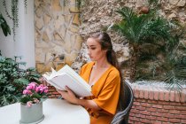 Вид збоку ніжна жінка читає цікаву книгу, сидячи за столом на літній терасі будинку — стокове фото
