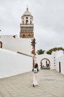 На міській вулиці міста Фуертевентура (Іспанія) перед спиною постає анонімна жінка з рюкзаком, що йде по тротуару проти білих будинків і похмурого сірого неба. — стокове фото