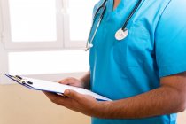 Medico maschio concentrato anonimo con stetoscopio leggere rapporto medico sugli appunti mentre in piedi in clinica — Foto stock