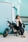 Motociclista feminina confiante apoiando-se na motocicleta estacionada na beira da estrada na cidade e fumar cigarro enquanto olha para longe — Fotografia de Stock
