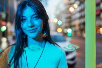 Joven hembra suave mirando a la cámara contra la calle urbana con luz azul artificial - foto de stock