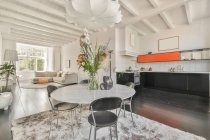Дизайн інтер'єру обідньої зони з круглим столом та стільцями, розміщеними на килимі у просторій квартирі з відкритим плануванням кухні та білих стін та стелі в сучасному стилі лофт — стокове фото
