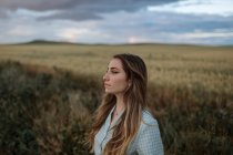 Side view of young mindful female looking away on road near meadow under cloudy sky in evening in countryside — Stock Photo