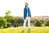 Positive junge Frau in trendiger Kleidung steht auf einem grünen Hügel vor dem Hintergrund der Stadt an einem sonnigen Tag und blickt in die Kamera im Gegenlicht — Stockfoto