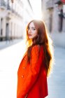 Вид сбоку стильной женщины с имбирными волосами и в ярко-оранжевом костюме, идущей по городской улице, глядя в камеру — стоковое фото