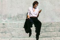 Donna deliziata in camicia bianca formale e pantaloni seduti sul bordo di cemento e avendo videochiamata — Foto stock