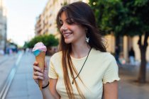 Colheita alegre jovem fêmea em pingente e brincos com delicioso gelato em cone de waffle olhando para longe na rua — Fotografia de Stock